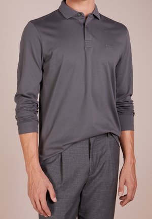 Man die een donkergrijze polo met lange mouwen en grijze broek draagt, ontspannen met zijn armen staand tegen een effen beige achtergrond.