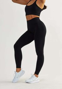 Soutien-gorge de sport noir et leggings taille haute en tissu extensible. Le modèle porte des chaussures de sport blanches avec des accents gris. Design lisse et ajusté.