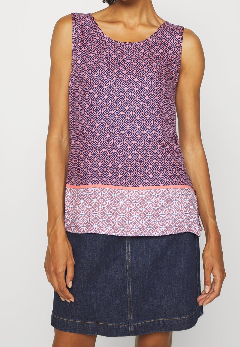 Mujer con blusa sin mangas de estampado morado y rosa y falda de mezclilla azul sobre un fondo liso.