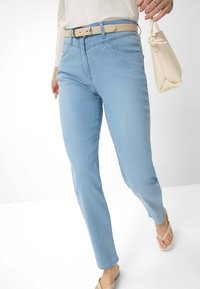 Lys blå jeans med slim fit, der har et beige bælte og en glat tekstur. En cremefarvet håndtaske holdes i højre hånd.