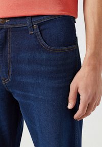 Jeans de mezclilla azul oscuro con costuras naranjas, diseño clásico de cinco bolsillos y un corte ajustado, con una vista cercana de la zona de la cintura.