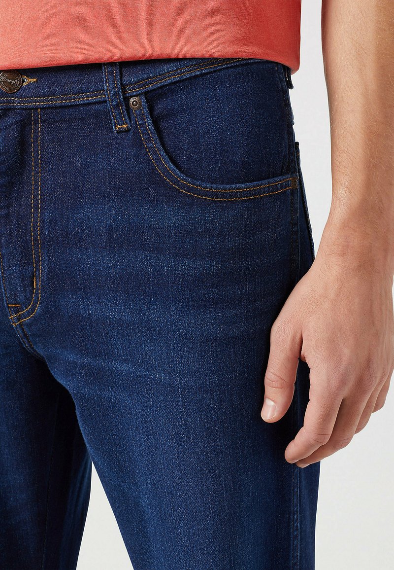 Granatowe dżinsy z pomarańczowym przeszyciem, klasyczny pięciopocketowy design i slim fit, z widokiem z bliska na strefę talii.