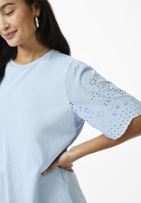 Lyseblå bomullstop med korte, broderte ermer som har blomster utskjæringer og bølgete kanter. Rund halslinje og avslappet passform.