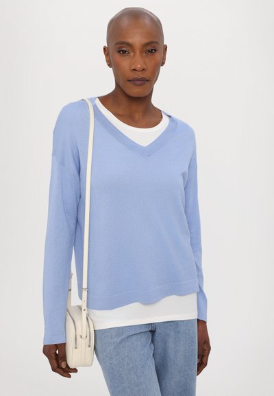 Light blue V-neck sweater layered over a white top, featured with long sleeves and a relaxed fit, paired with light blue jeans and a small white bag.