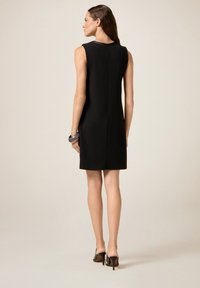 Robe noire sans manches à coupe droite, dotée d'une fermeture éclair discrète au dos et d'un tissu lisse. Accentuée par des chaussures à talons hauts noires.