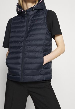 Person trägt eine marineblaue gesteppte ärmellose Daunenweste mit Kapuze über einem schwarzen kurzärmeligen Shirt und schwarzen Hosen.