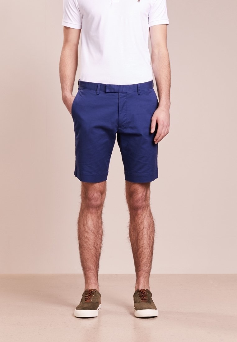 Shorts pour hommes en tissu de coton bleu, avec poches latérales, devant plat et ceinture standard. Associés à des baskets vert olive.