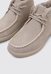 Botas de tobillo de ante beige con forro de piel sintética, puntera redondeada, costuras decorativas y suela de goma plana. Detalle de cordones y logo visible.