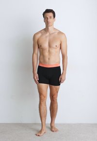 Zwarte boxershorts met een feloranje tailleband. Het materiaal lijkt rekbaar, getailleerd en heeft een gladde textuur. Het model staat op blote voeten.