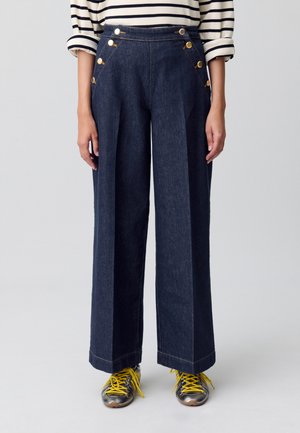 Pantalones de mezclilla oscuros de pierna ancha con botones dorados en la parte frontal. Presentan doble pinza y una cinturilla estructurada. Combinados con calzado informal.