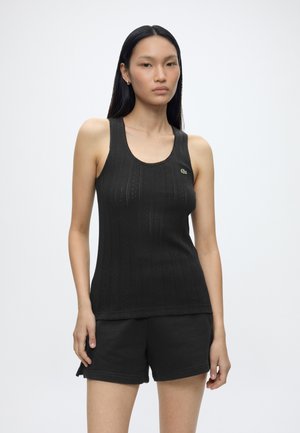Femme aux longs cheveux noirs portant un débardeur sans manches noir texturé et un short noir, debout devant un fond clair uni.
