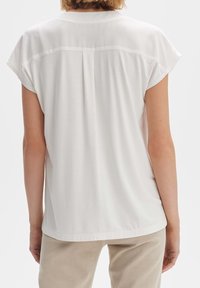Weiße Bluse mit kurzen Ärmeln, runder Ausschnitt und gefaltetem Rückendetail. Weicher, leichter Stoff fällt sanft. Saubere Nähte und ein schlichtes Design.