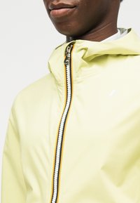 Gros plan sur une personne portant une veste à capuche jaune pâle avec une fermeture éclair noire, blanche et orange, montrant le bas du visage et le cou.