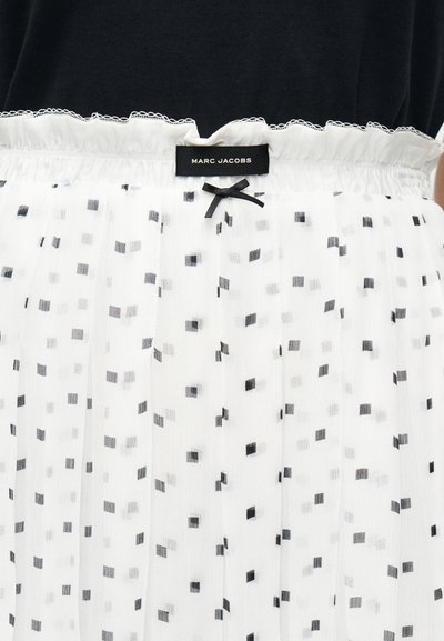 Λευκό ύφασμα με μικρό μοτίβο μαύρων τετραγώνων, ελαστική μέση με μαζεμένο ύφασμα και μαύρο φιόγκο, και μαύρη ετικέτα ρούχων με την επιγραφή "Marc Jacobs" στην κορυφή.