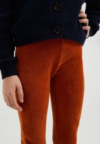 Mornarsko modra pletena kardigan z debelimi pleteninami in tremi velikimi gumbi, v kombinaciji s pomarančnimi žametnimi hlačami z rebrasto teksturo, ki prikazujejo vzorec vertikalnih črt.