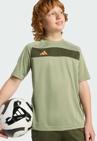 Svetle zelené sportovní tričko s krátkými rukávy, s tmavě zeleným vodorovným pruhem a oranžovým logem. Drží se černobílý fotbalový míč.