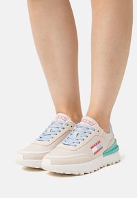 Zapatillas beige con cordones azul claro y acentos verdes. Presentan una parte superior texturizada, suela blanca y detalles del logo rojo en el costado.