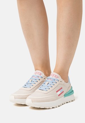 Zapatillas beige con cordones azul claro y acentos verdes. Presentan una parte superior texturizada, suela blanca y detalles del logo rojo en el costado.