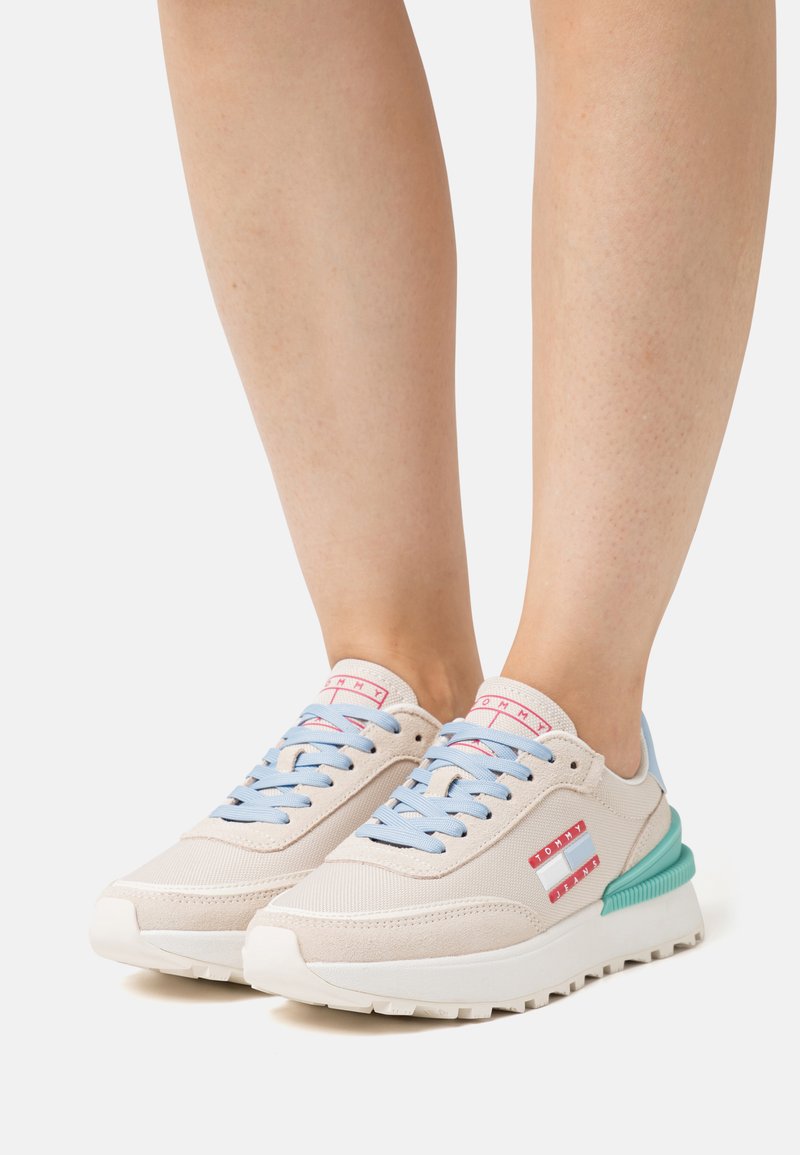 Zapatillas beige con cordones azul claro y acentos verdes. Presentan una parte superior texturizada, suela blanca y detalles del logo rojo en el costado.