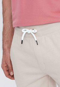 Pantalons de survêtement beige clair avec une taille côtelée et un cordon blanc. Le tissu semble doux avec des détails de couture visibles.