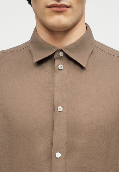 Άνδρας που φοράει ένα ανοιχτό καφέ πουκάμισο με κουμπιά και κλασικό γιακά, εμφανιζόμενος από τους ώμους μέχρι το στόμα σε λευκό φόντο.