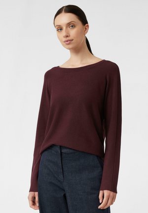Burgunderfarbener Langarmpullover mit weit ausgeschnittenem Hals und Rippdetails an den Ärmeln, kombiniert mit dunklen Denim-Hosen. Glatte Textur.