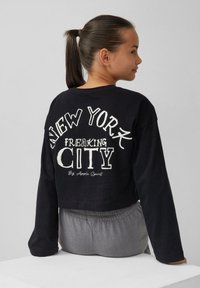 Zwarte cropped sweatshirt met lange mouwen, met gedurfde witte tekst "NEW YORK FREAKING CITY" op de achterkant en "Big Apple Spirit" eronder.