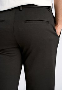 Zwarte broek van gladde stof, met een rechte snit, standaard zakken en een tailleband met lussen voor een riem. De achterkant toont minimale details.
