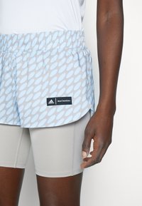 Atlet v modro-šedých vzorovaných šortkách Adidas x Marimekko přes světle šedé kompresní šortky, stojící na bílém pozadí.