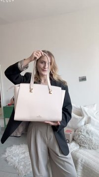 Ružová kožená kabelka na nosenie v ruke s dvoma hornými rukoväťami, zlatými kovovými doplnkami a štruktúrovaným tvarom, držená osobou v čiernom blejzri.