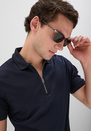 Giovane uomo con polo blu navy con cerniera che si sistema degli occhiali da sole neri sul viso davanti a uno sfondo semplice.
