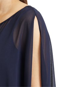 Gros plan sur une personne portant un chemisier transparent bleu marine avec une fente à l'épaule laissant apparaître la peau, sur fond blanc.