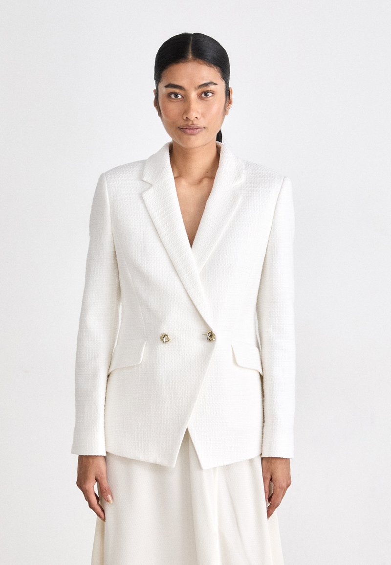 White double-breasted blazer in textured fabric, featuring notch lapels, gold-tone hardware, and two front pockets. Smooth, structured design.