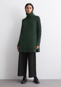 Grøn strikket turtleneck-sweater med struktureret mønster, sidelægninger og lange ærmer, parret med sorte vide bukser og sorte sko.