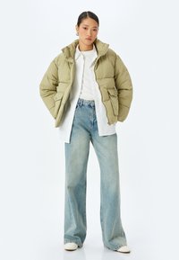 Giacca imbottita verde oliva con cappuccio, tasche frontali e design trapuntato, abbinata a una camicia bianca e jeans larghi a gamba larga azzurri.