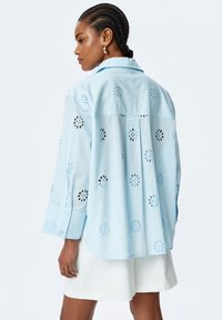 Hellblaues Hemd mit floralem Aussschnittmuster, langen Ärmeln und abgerundetem Saum. Hergestellt aus weichem Stoff. Lässiges Design.