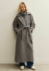 Abrigo largo de lana gris con cinturón, gran cuello y mangas abotonadas. Lleva puesto un suéter de cuello alto gris, combinado con jeans azules y zapatillas blancas.