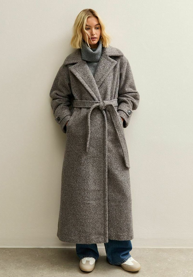 Abrigo largo de lana gris con cinturón, gran cuello y mangas abotonadas. Lleva puesto un suéter de cuello alto gris, combinado con jeans azules y zapatillas blancas.