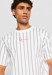 White cotton t-shirt with black vertical stripes, featuring a round neck and short sleeves. Red signature logo on the chest.