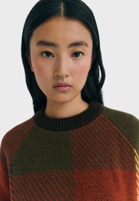 Una giovane donna con lunghi capelli scuri indossa un maglione lavorato a maglia con grandi motivi a scacchi verde e rosso, posando frontalmente su uno sfondo semplice.