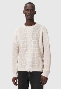 Krémový sveter s pleteným vzorom, ktorý obsahuje kombináciu textúrovaných vzorov, žebrované manžety a jemne rozstrapkaný lem. Nosené s tmavými džínami.