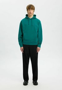 Felpa verde in cotone con tasca a marsupio e cappuccio regolabile, abbinata a pantaloni neri e scarpe nere su uno sfondo neutro.