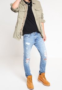Persona vestida con una chaqueta color marrón claro, camisa negra, jeans rasgados de color azul claro, y botas de cordones marrón claro, posando con una mano cerca de su cabeza.