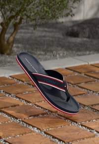 Black flip-flop with red and white striped straps, featuring a brand logo. Textured sole, lightweight design, and elevated position above a wooden surface.