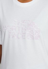 Bílá tričko s krátkým rukávem, které má na hrudi umístěné světle fialové logo s texturovaným vzorem, kulatý výstřih a měkkou látku.