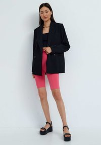 Blazer nero oversize con doppi bottoni, indossato sopra un crop top nero e pantaloni shorts in ciclismo rosa; abbinato a sandali platform neri chunky.