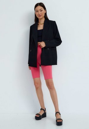 Musta ylipitkä bleiseri, jossa on kaksoispainikkeet, päällä musta crop-top ja vaaleanpunaiset pyöräilyshortsit; yhdistettynä chunky-mustiin platform-sandaaleihin.