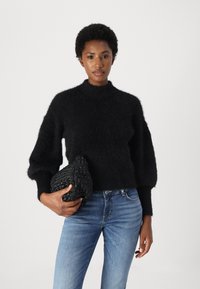 Maglione nero, soffice, a maglia con collo alto e maniche a palloncino, abbinato a jeans blu e a una borsa nera con texture.