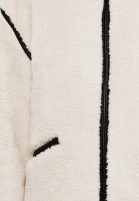 Μπουφάν από φλις σε εκρού χρώμα με υφή, που διαθέτει κεντρικό μαύρο φερμουάρ και πλαϊνή τσέπη με μαύρο περίγραμμα.