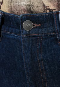 Los vaqueros de mezclilla azul oscuro presentan un botón de metal con un símbolo de infinito, costuras marrones y una vista en primer plano de la cinturilla.